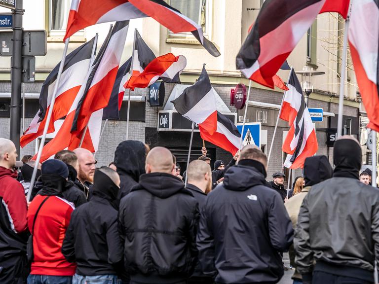 Mehrere Teilnehmer einer Demonstration der Neonazi-Gruppe "Jung und Stark" stehen mit szenetypischen Kleidungstücken nebeneinander. Im Hintergrund sind schwarz-weiß-rote Fahnen zu sehen.