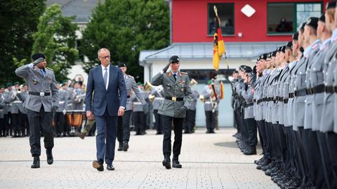 Bundeskanzler Merz geht zwischen zwei Bundeswehrsoldaten an einer Reihe von uniformierten Rekruten vorbei.