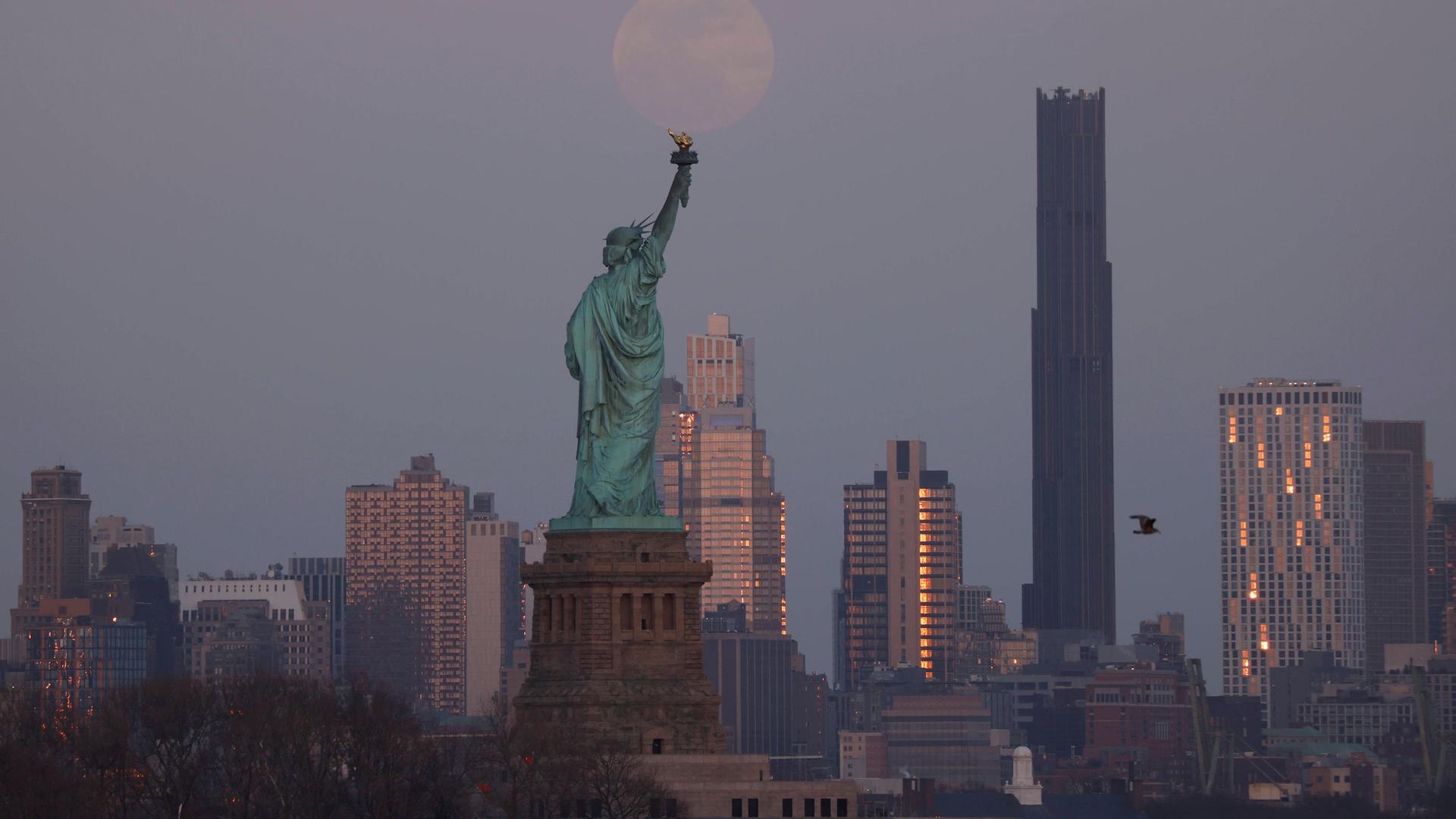 Auf einer Aufnahme ist die Freiheitsstatue von hinten zu sehen. Im Hintergrund sind New Yorker Wolkenkratzer und der Mond zu erkennen. Auf einer Aufnahme ist die Freiheitsstatue von hinten zu sehen. Im Hintergrund sind New Yorker Wolkenkratzer und der Mond zu erkennen.