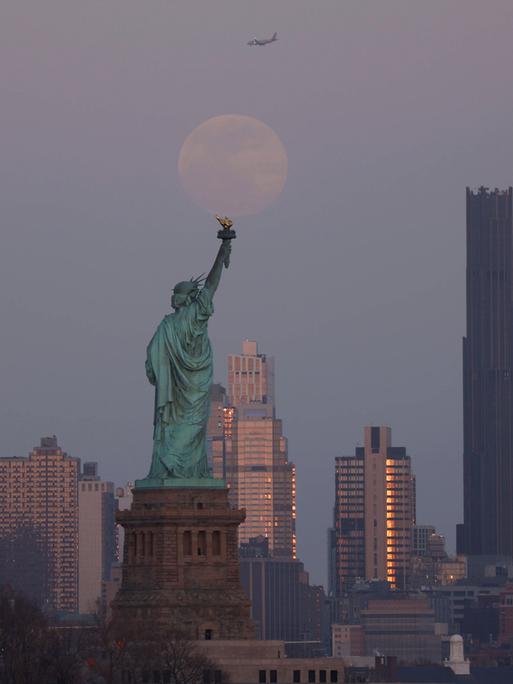 Auf einer Aufnahme ist die Freiheitsstatue von hinten zu sehen. Im Hintergrund sind New Yorker Wolkenkratzer und der Mond zu erkennen.
