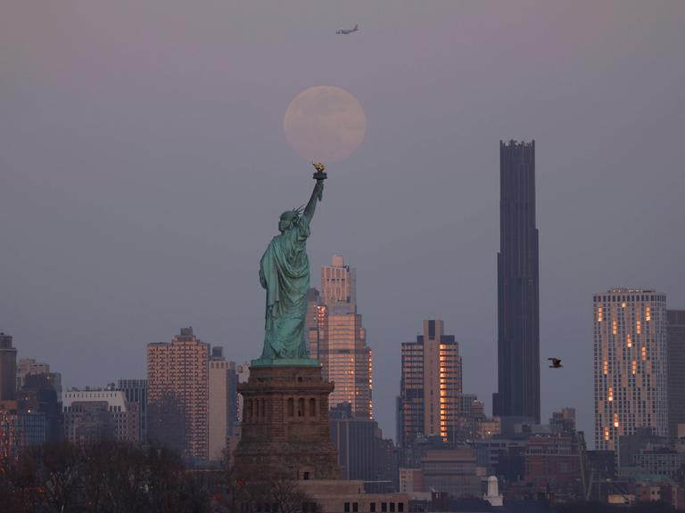 Auf einer Aufnahme ist die Freiheitsstatue von hinten zu sehen. Im Hintergrund sind New Yorker Wolkenkratzer und der Mond zu erkennen.