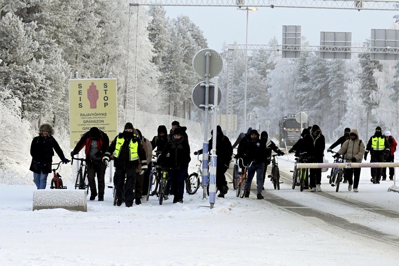 Finnische Grenzschutzbeamten eskortieren Migranten mit Fahrrädern am internationalen Grenzübergang in Salla. Ringsherum ein verschneiter Wald. 