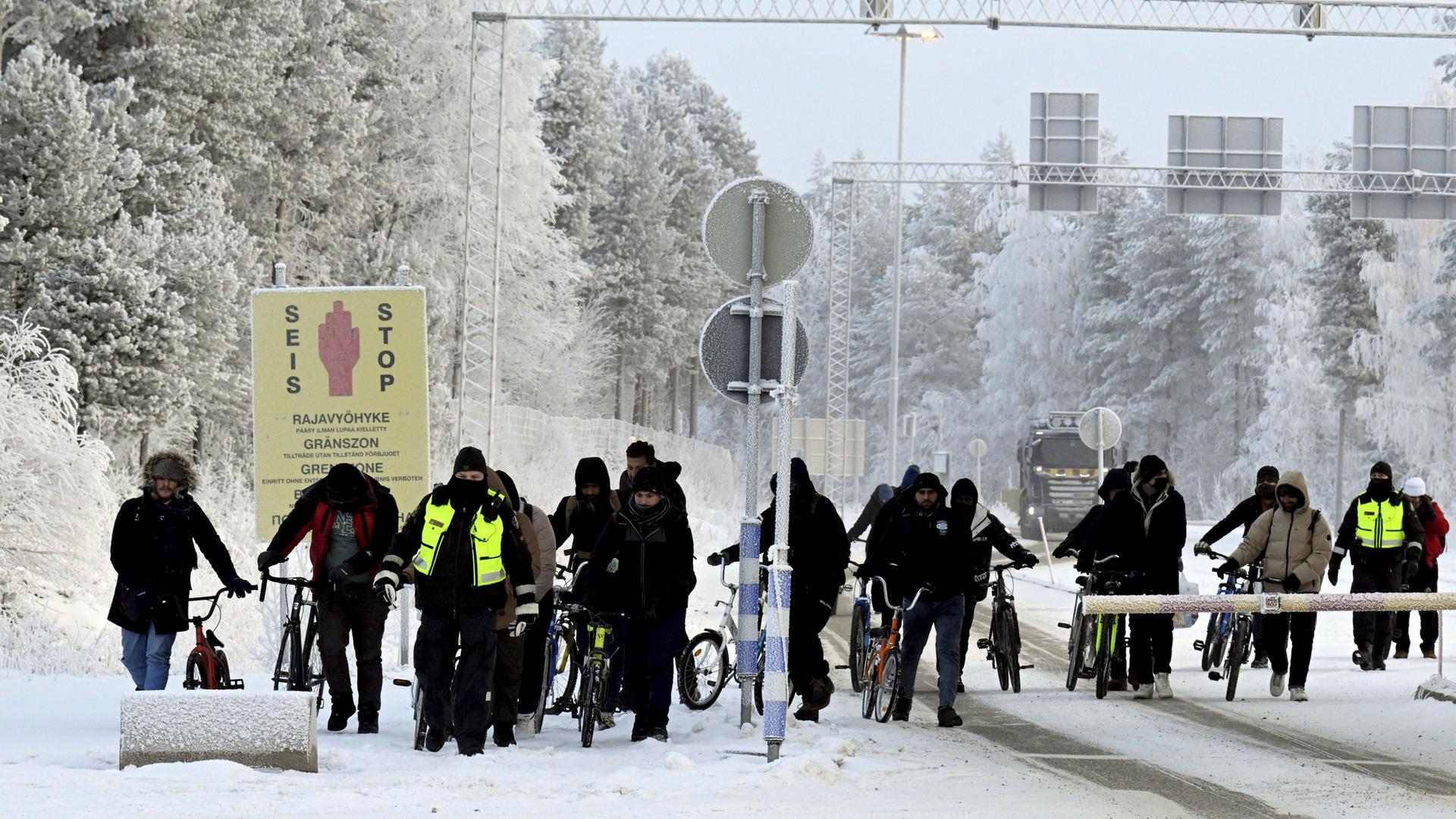 Finnische Grenzschutzbeamten eskortieren Migranten mit Fahrrädern am internationalen Grenzübergang in Salla. Ringsherum ein verschneiter Wald.