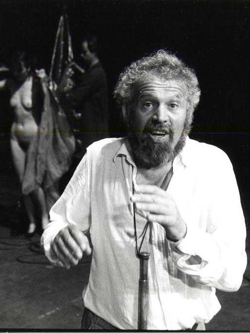 Otto M. Zykan auf der Bühne mit zwei Darstellern, darunter eine nackte Frau, im Hintergrund