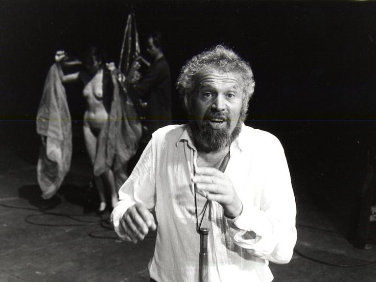 Otto M. Zykan auf der Bühne mit zwei Darstellern, darunter eine nackte Frau, im Hintergrund