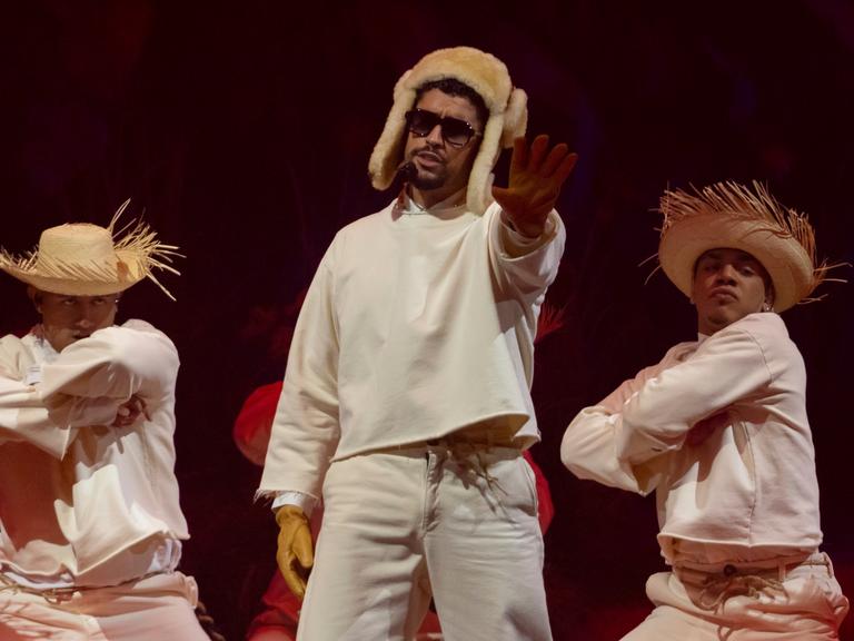 Der Latino-Rapper Bad Bunny trägt eine russische Fellmütze und steht mit zwei Tänzern mit Strohhut auf der Bühne in seiner Heimat Puerto Rico
