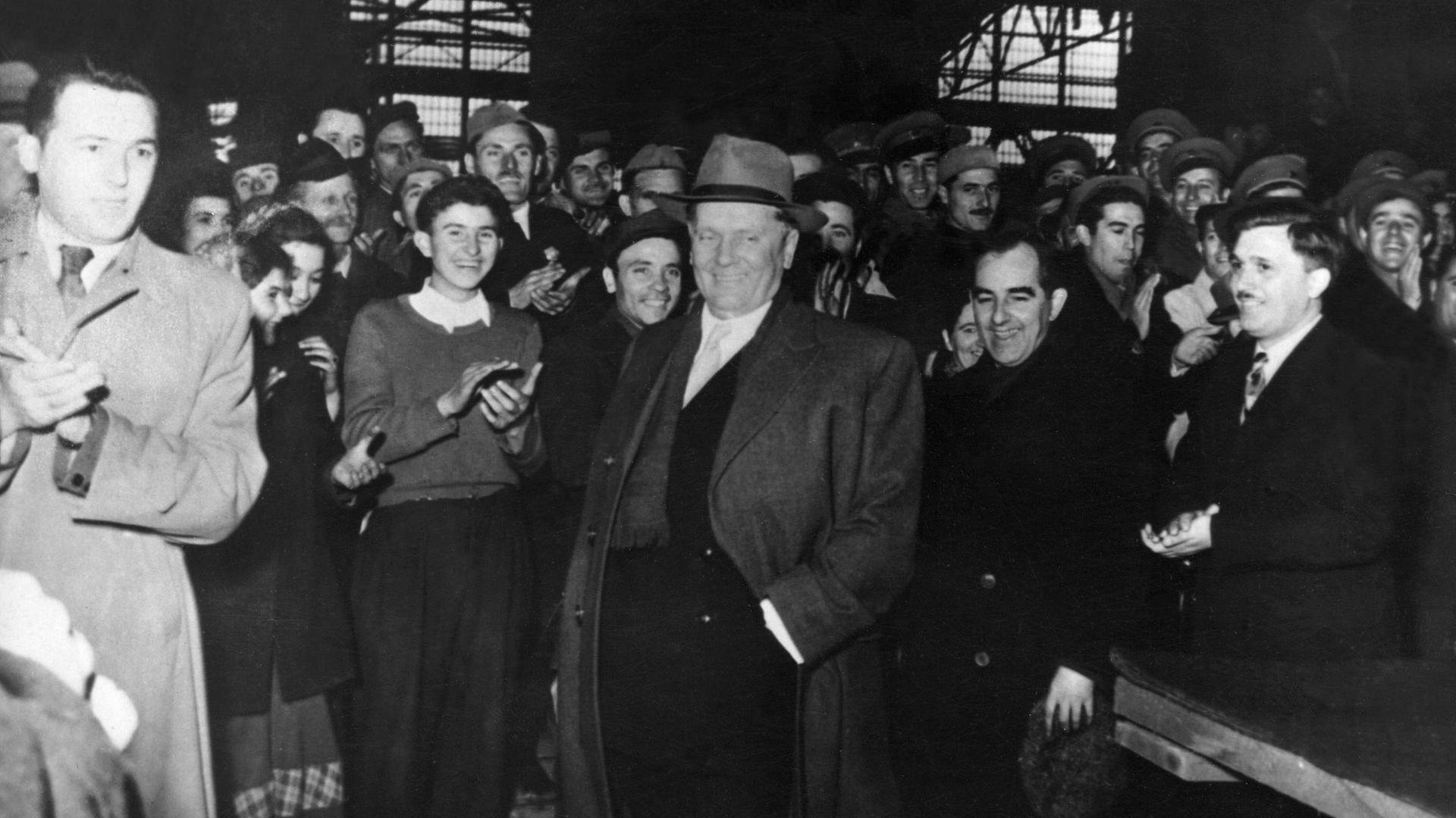 Schwarz-weiß Foto eines Besuchs von Josip Tito in einer jugoslawischen Fabrik in den 1950er Jahren. Er trägt Mantel und Hut. Um ihn herum stehen Fabrikarbeiter und klatschen.