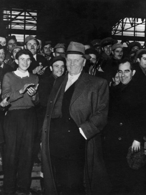 Schwarz-weiß Foto eines Besuchs von Josip Tito in einer jugoslawischen Fabrik in den 1950er Jahren. Er trägt Mantel und Hut. Um ihn herum stehen Fabrikarbeiter und klatschen.