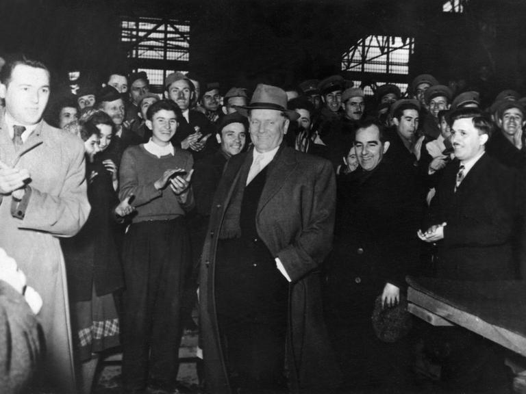 Schwarz-weiß Foto eines Besuchs von Josip Tito in einer jugoslawischen Fabrik in den 1950er Jahren. Er trägt Mantel und Hut. Um ihn herum stehen Fabrikarbeiter und klatschen.