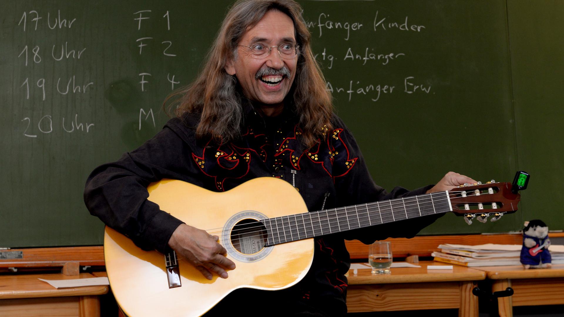 Peter Bursch steht mit einer Gitarre in Händen vor einer Tafel. 