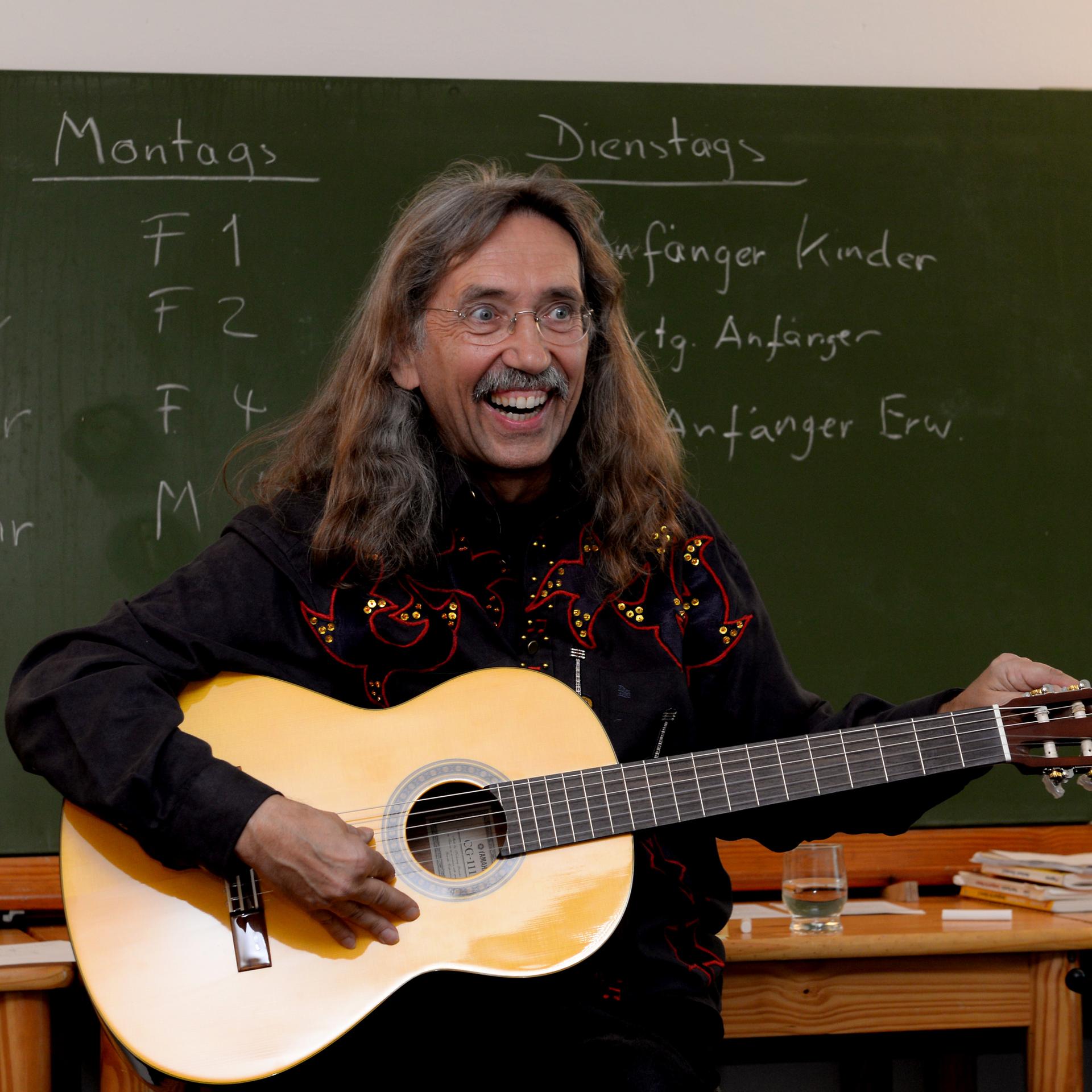 Peter Bursch steht mit einer Gitarre in Händen vor einer Tafel. 