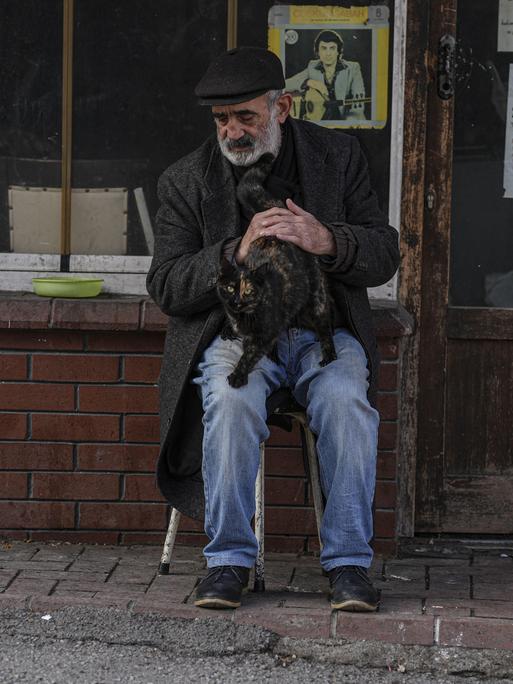 Ein älterer Gebrauchtwarenhändler sitzt in Ankara vor seinem kleinen Laden und streichelt eine Katze auf seinem Schoß.
