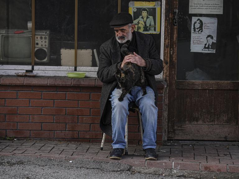 Ein älterer Gebrauchtwarenhändler sitzt in Ankara vor seinem kleinen Laden und streichelt eine Katze auf seinem Schoß.