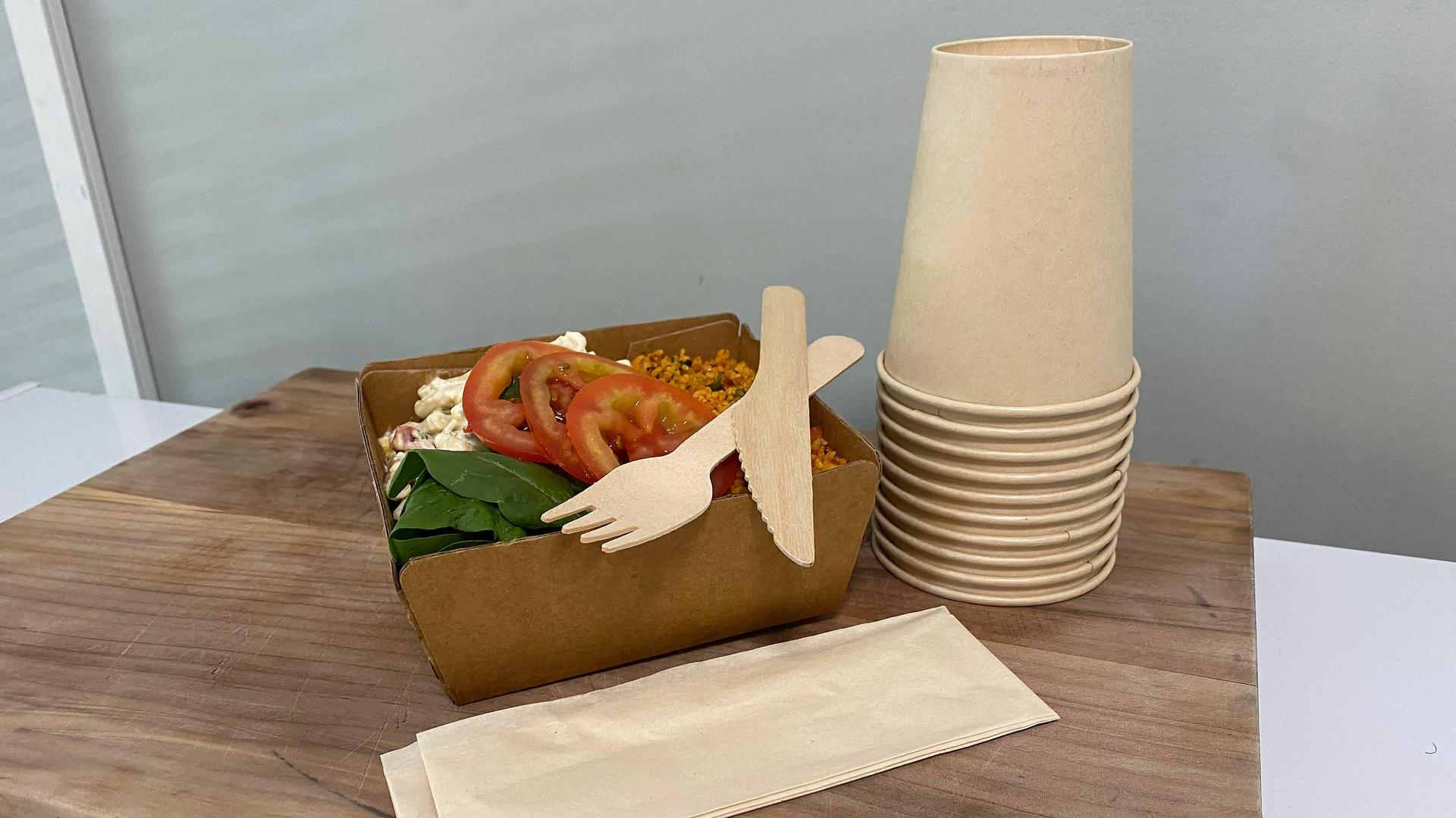 In einem Pappteller befindet sich ein Salat, darauf liegen eine Gabel und ein Messer aus Holz, daneben steht ein Stapel Pappbecher.