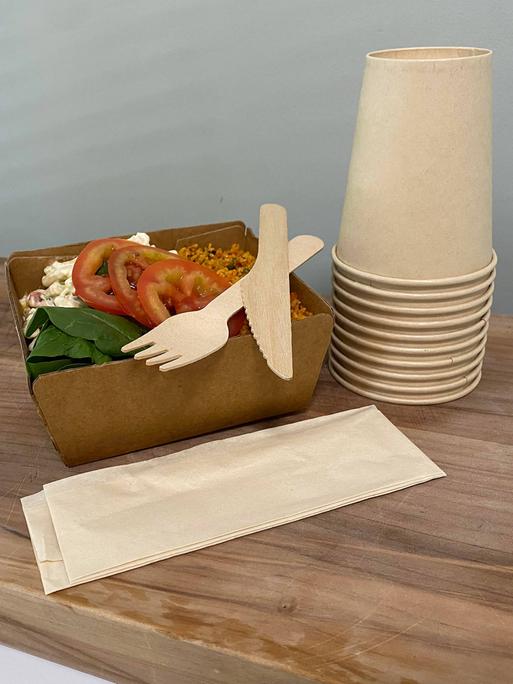 In einem Pappteller befindet sich ein Salat, darauf liegen eine Gabel und ein Messer aus Holz, daneben steht ein Stapel Pappbecher.