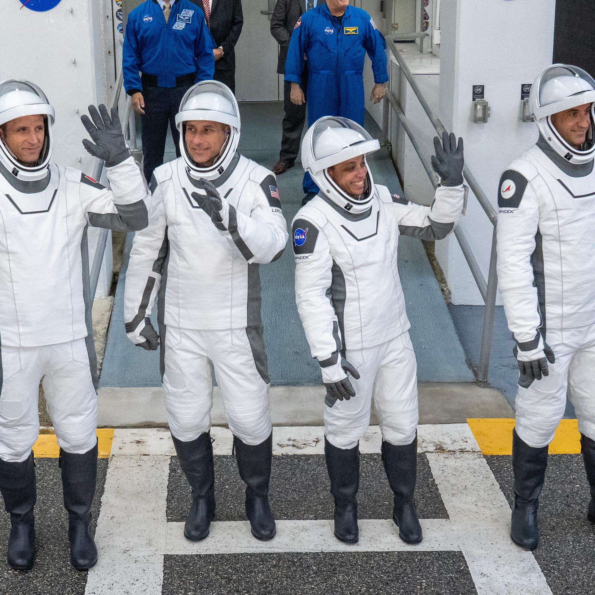 Das Bild zeigt die NASA-Astronautin Jessica Watkins und ihre Kollegen Luke Delaney, Joshua Kutryk und Sergey Teteryatnikov in weißen Raumanzügen mit Helm. Sie winken fröhlich in die Menge.
