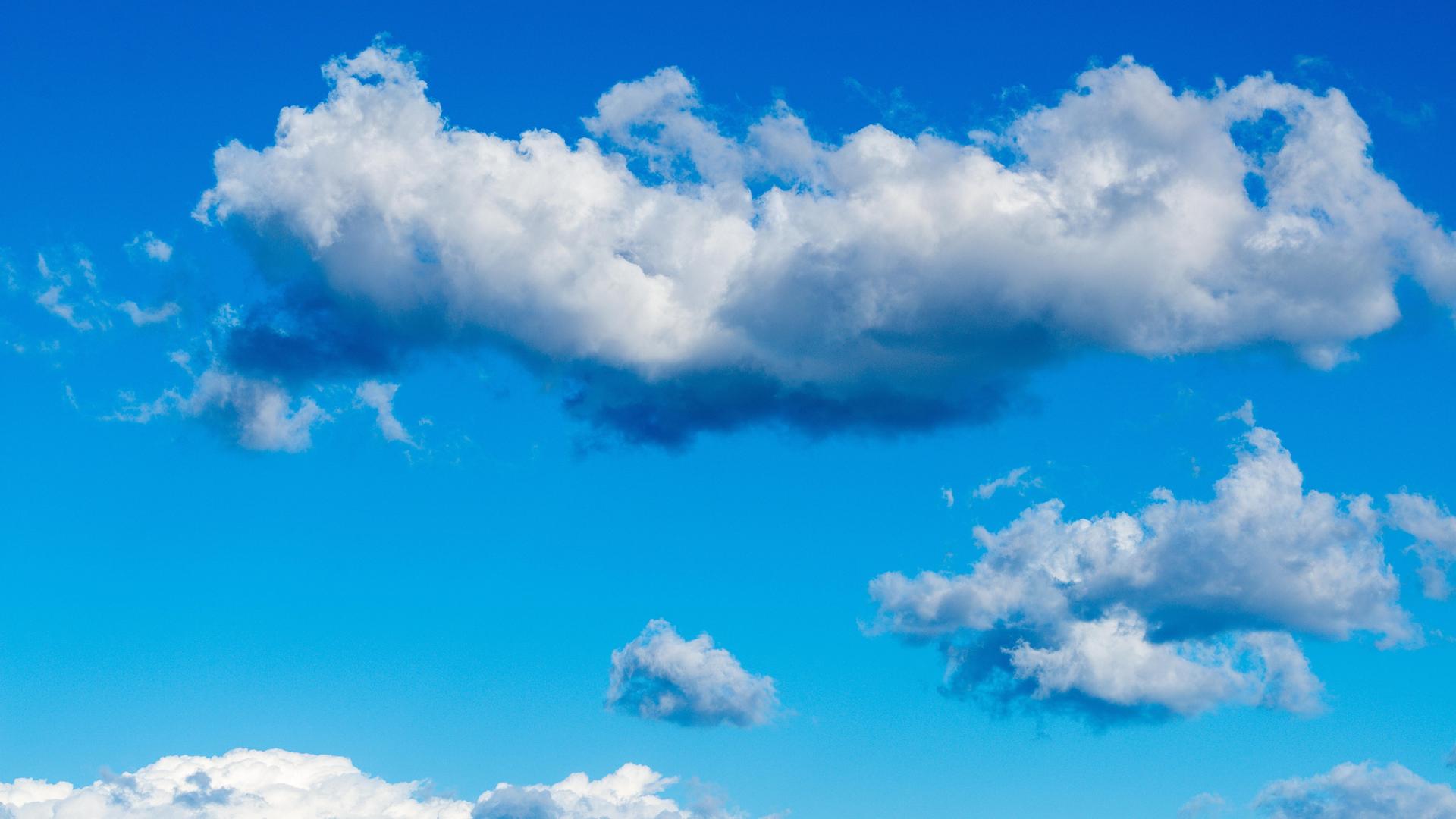 Hellblauer Himmel ist mit weichen kumulierten Wolkenformen gefüllt, darunter ein langes dichtes Wolkenband und verstreut kleinere Wolken in der Entfernung. 