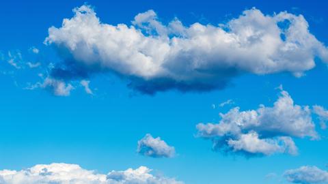 Hellblauer Himmel ist mit weichen kumulierten Wolkenformen gefüllt, darunter ein langes dichtes Wolkenband und verstreut kleinere Wolken in der Entfernung. 