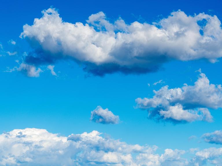 Hellblauer Himmel ist mit weichen kumulierten Wolkenformen gefüllt, darunter ein langes dichtes Wolkenband und verstreut kleinere Wolken in der Entfernung. 