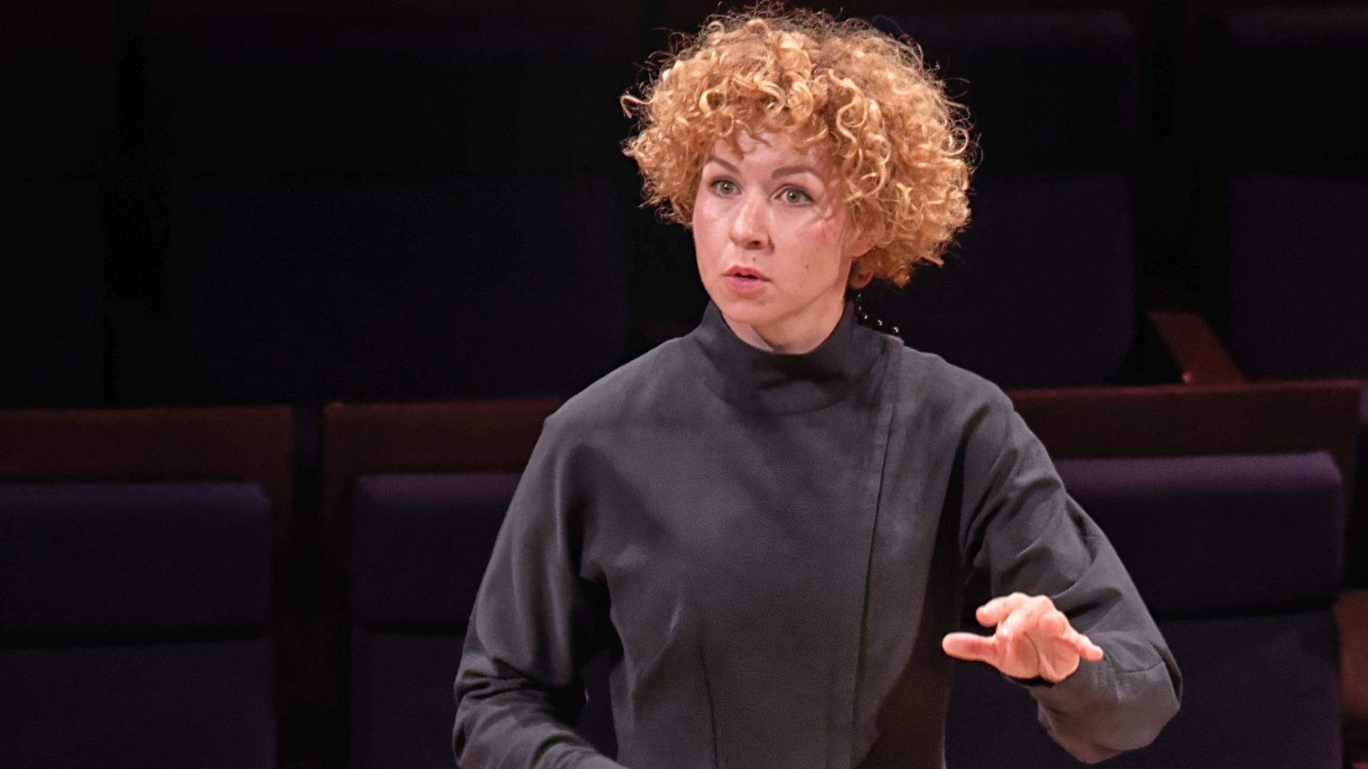 Bild einer jungen Frau mit blonden Locken, die ein schwarzes Kleid trägt und einen Dirigentenstab in der Hand hält Bild einer jungen Frau mit blonden Locken, die ein schwarzes Kleid trägt und einen Dirigentenstab in der Hand hält