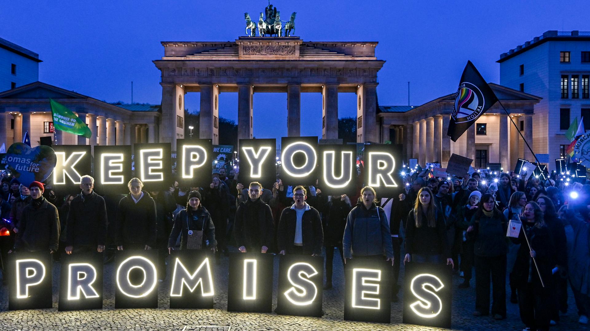 Mehrere Menschen halten in der Dämmerung leuchtende Buchstaben vor dem Brandenburger Tor in Berlin hoch. Daraus bildet sich der Schriftzug "Keep your promises".
