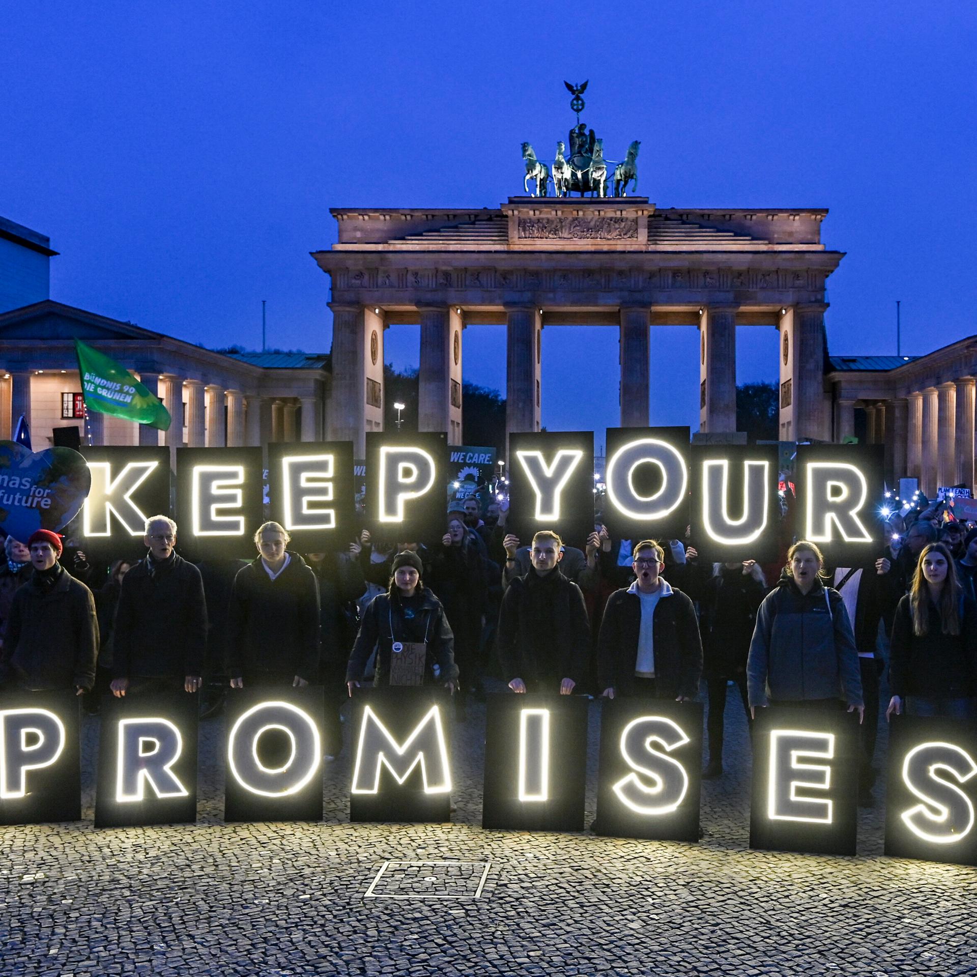 Mehrere Menschen halten in der Dämmerung leuchtende Buchstaben vor dem Brandenburger Tor in Berlin hoch. Daraus bildet sich der Schriftzug "Keep your promises".