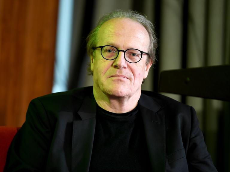 Thomas Flierl schaut direkt in die Kamera. Er trägt ein schwarzes Sakko und eine runde schwarze Brille.