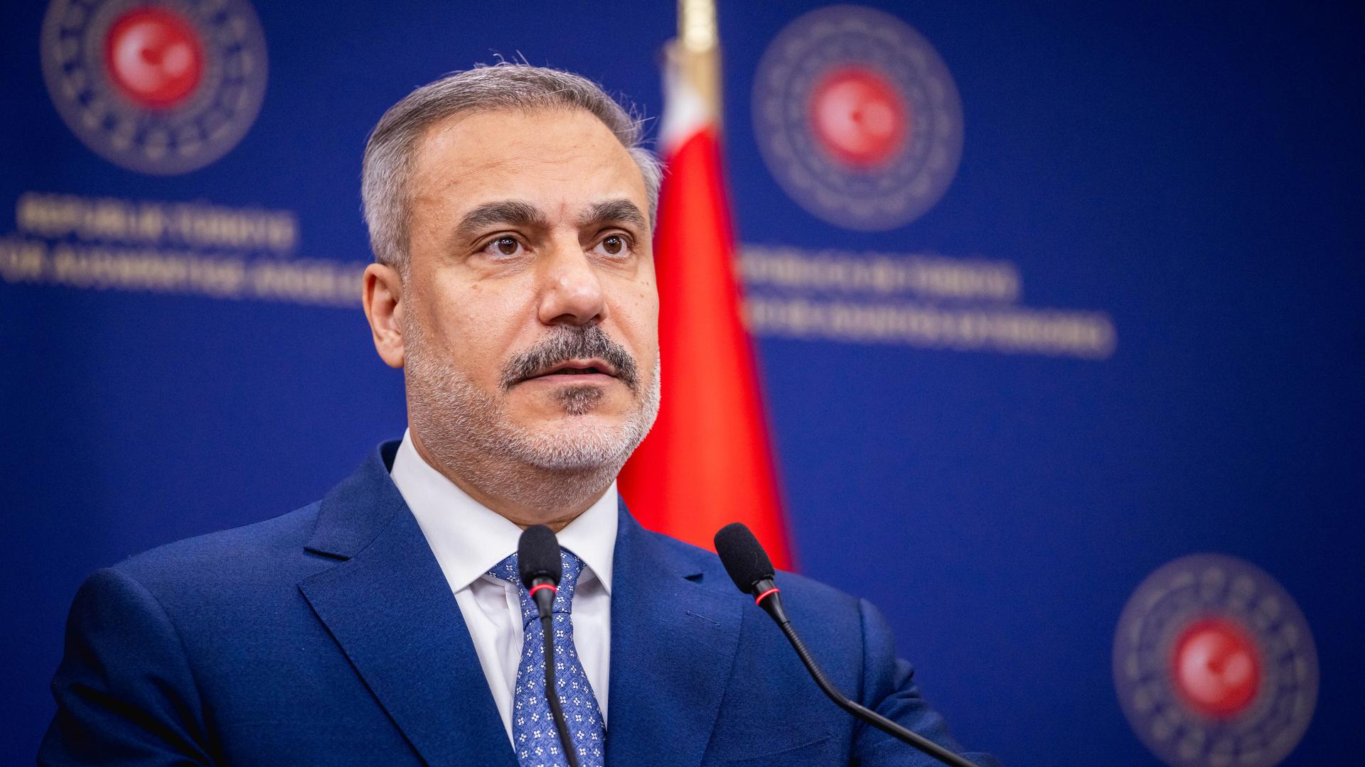 Porträtaufnahme des türkischen Außenministers Hakan Fidan. Er trägt einen Anzug und spricht auf einem Podium in ein Mikrofon.