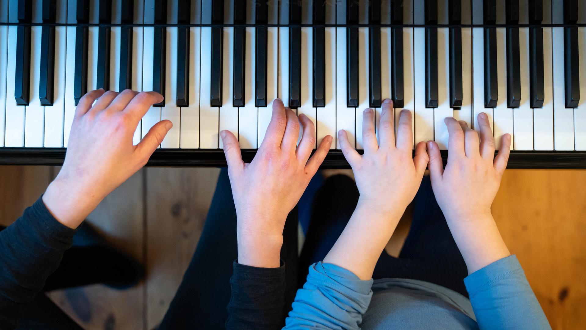 Zwei Paar Hände  liegen auf einer Klaviertastatur: Eine Frau bringt einem Mädchen Klavierspielen bei. 