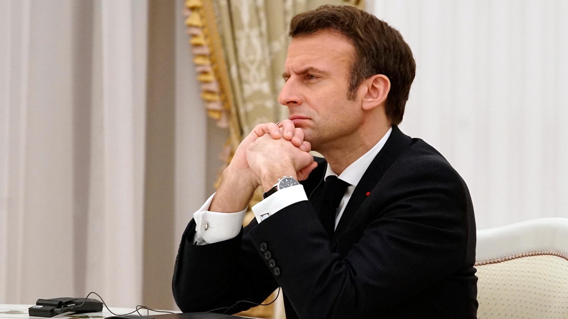Der französische Präsident Emmanuel Macron sitzt mit ernstem Blick, den Kopf auf die gefalteten Hände gestützt, an einem Verhandlungstisch im Kreml. Hinter ihm: die goldene Borte eines Vorhangs.