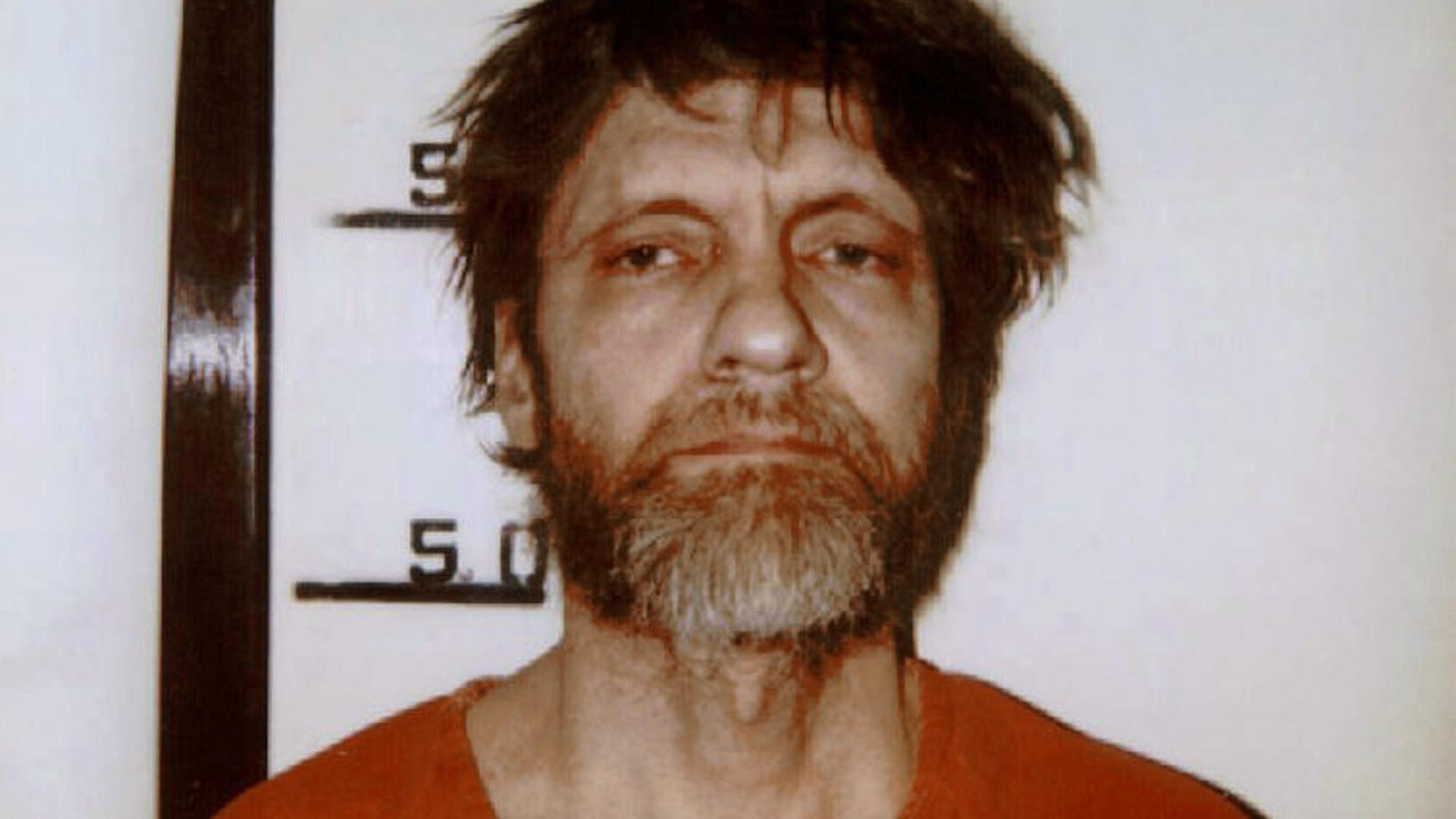 Mugshot von Ted Kaczynski in seinem orangeroten Gefängnisoverall. Sein Haar ist strubbelig und er schaut müde in die Kamera.