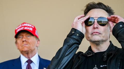 Musk setzt sich eine verspiegelte, schwarze Sonnenbrille auf und schaut in die Kamera. Im Hintergrund steht Donald Trump.