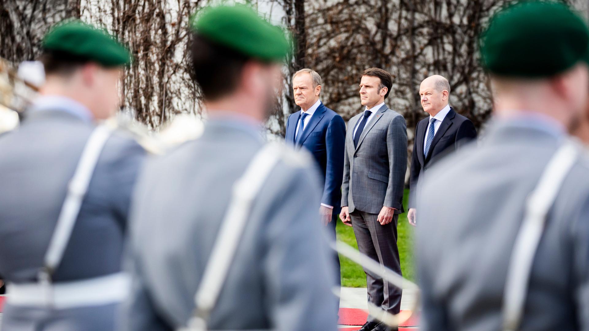 Bundeskanzler Olaf Scholz (SPD) steht neben Emmanuel Macron, Präsident von Frankreich, und Donald Tusk, Ministerpräsident von Polen. Im Vordergrund sind mehrere Soldaten in Uniform zu sehen, die die Gäste mit militärischen Ehren empfangen.