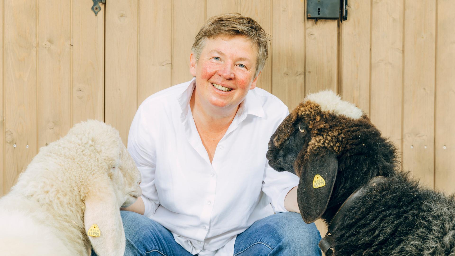 Porträt von Carolin Feismann mit den Schafen Alba und Pippa auf dem Gut Feismann, vor einer Holztür. Carolin Feismann guckt in die Kamera. Porträt von Carolin Feismann mit den Schafen Alba und Pippa auf dem Gut Feismann, vor einer Holztür. Carolin Feismann guckt in die Kamera.