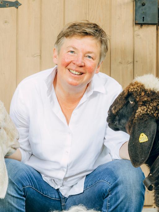 Porträt von Carolin Feismann mit den Schafen Alba und Pippa auf dem Gut Feismann, vor einer Holztür. Carolin Feismann guckt in die Kamera.