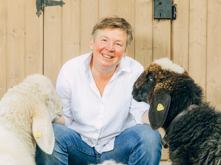 Porträt von Carolin Feismann mit den Schafen Alba und Pippa auf dem Gut Feismann, vor einer Holztür. Carolin Feismann guckt in die Kamera.