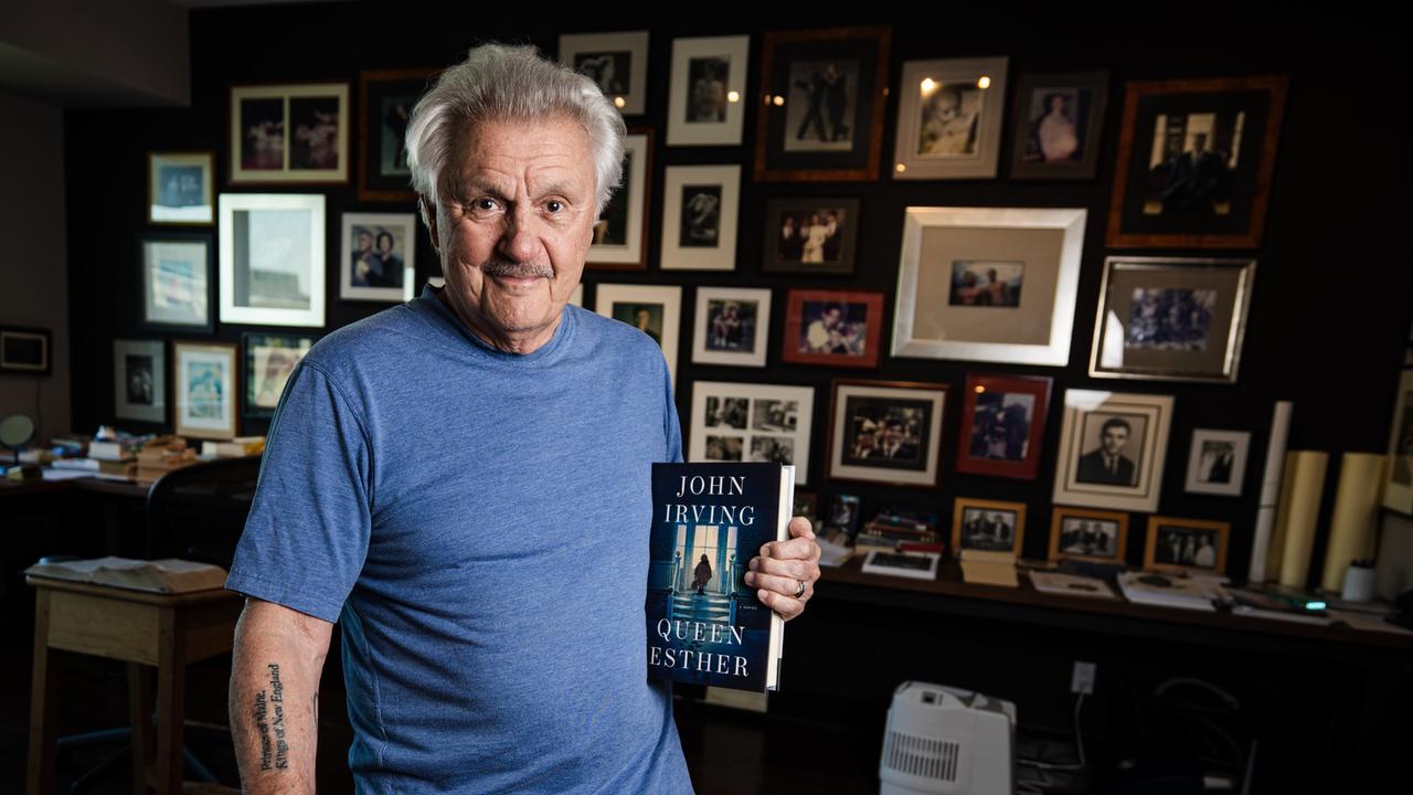 John Irving steht in seinem Arbeitszimmer in Toronto mit dem Buch "Queen Esther" in der Hand. Im Hintergrund ist eine Wand zu sehen, die über und über mit Bilderrahmen gefüllt ist.