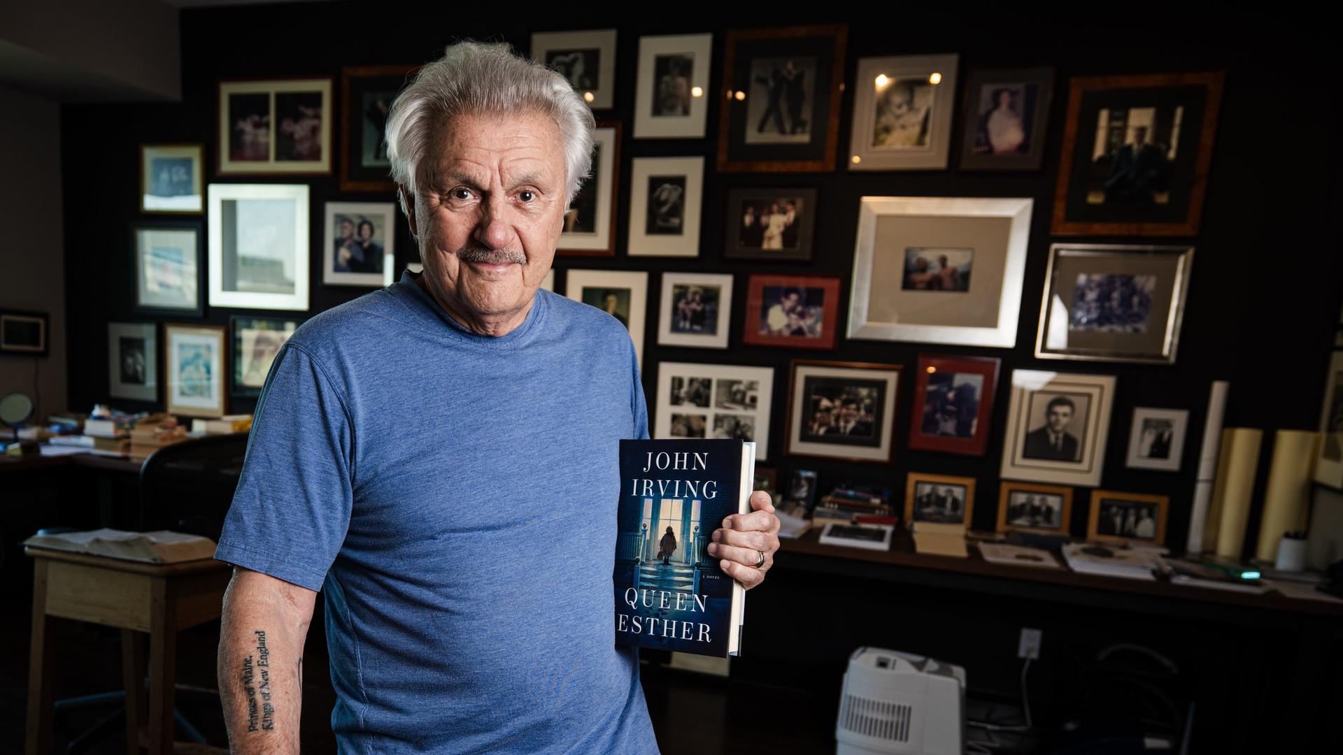 John Irving steht in seinem Arbeitszimmer in Toronto mit dem Buch "Queen Esther" in der Hand. Im Hintergrund ist eine Wand zu sehen, die über und über mit Bilderrahmen gefüllt ist. John Irving steht in seinem Arbeitszimmer in Toronto mit dem Buch "Queen Esther" in der Hand. Im Hintergrund ist eine Wand zu sehen, die über und über mit Bilderrahmen gefüllt ist.