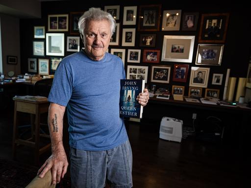 John Irving steht in seinem Arbeitszimmer in Toronto mit dem Buch "Queen Esther" in der Hand. Im Hintergrund ist eine Wand zu sehen, die über und über mit Bilderrahmen gefüllt ist.