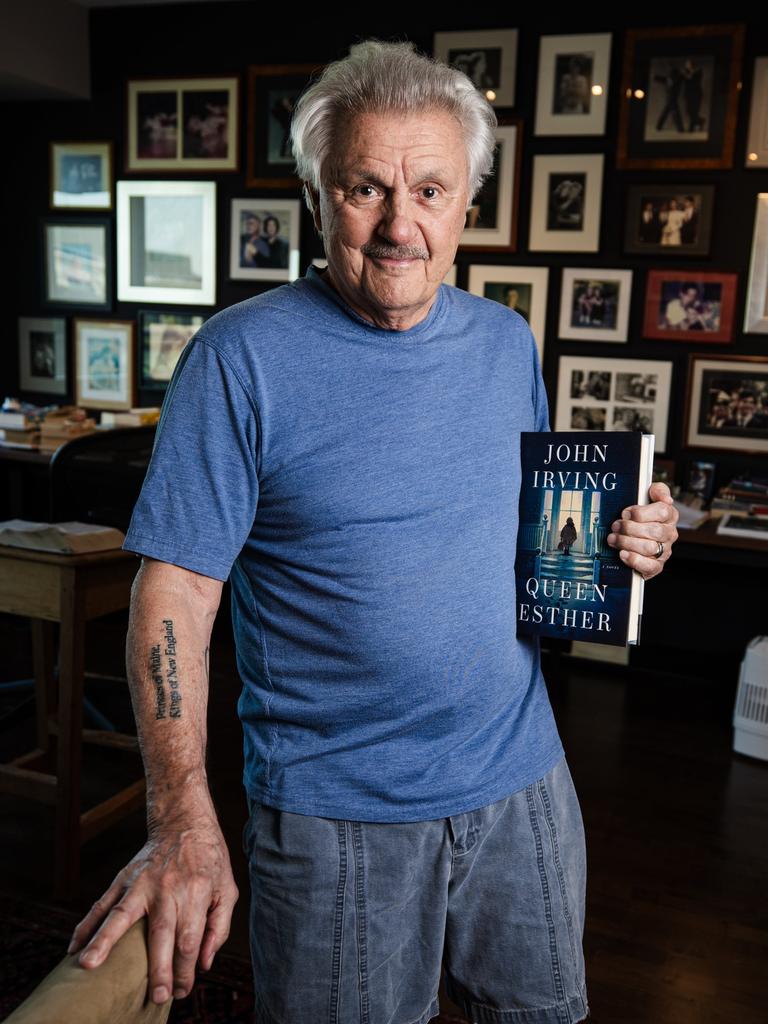John Irving steht in seinem Arbeitszimmer in Toronto mit dem Buch "Queen Esther" in der Hand. Im Hintergrund ist eine Wand zu sehen, die über und über mit Bilderrahmen gefüllt ist.