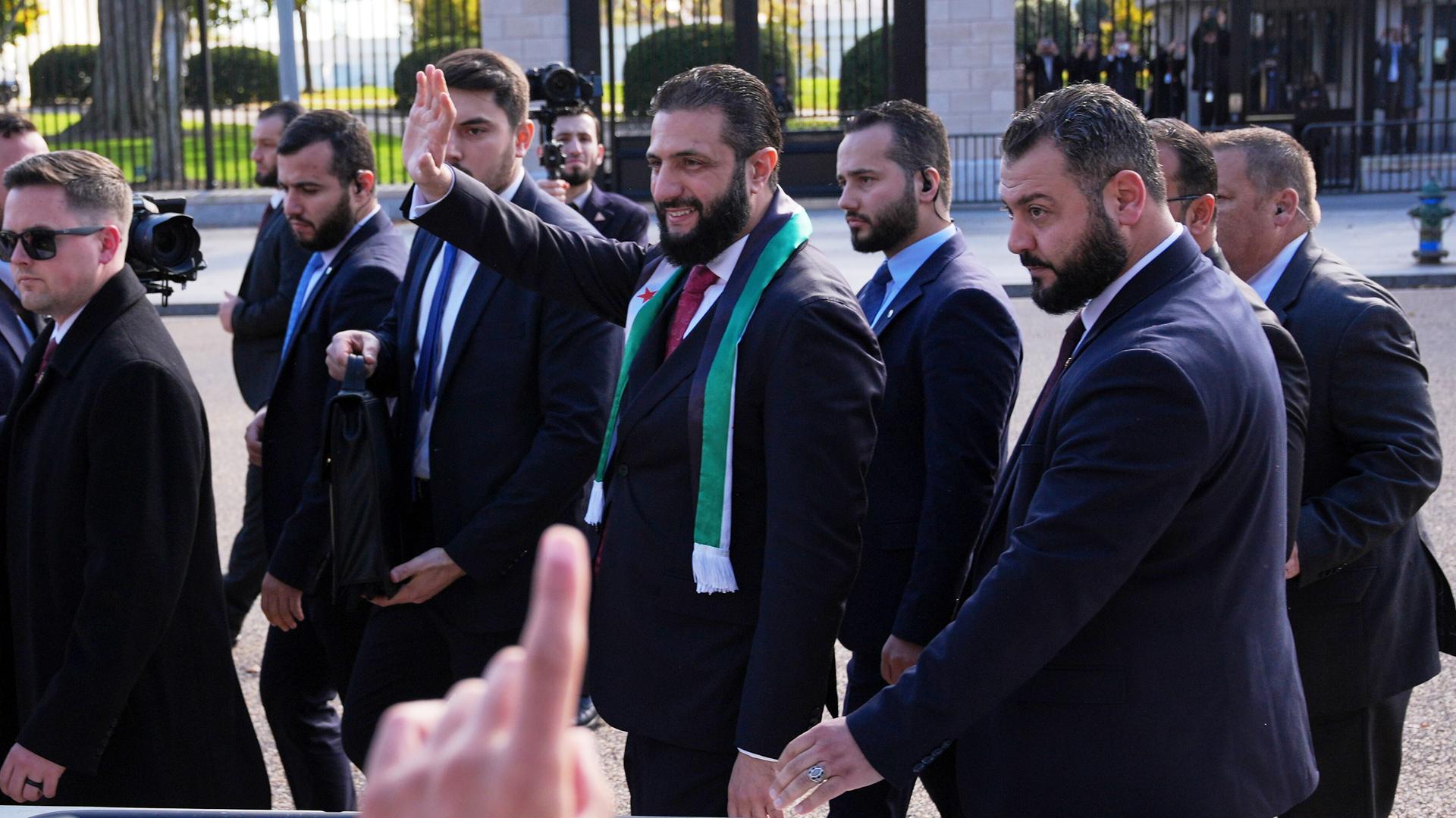 Der syrische Übergangspräsident al Scharaa winkt mit seiner Hand Menschen zu. Er ist von mehreren Sicherheitspersonen umgeben. 