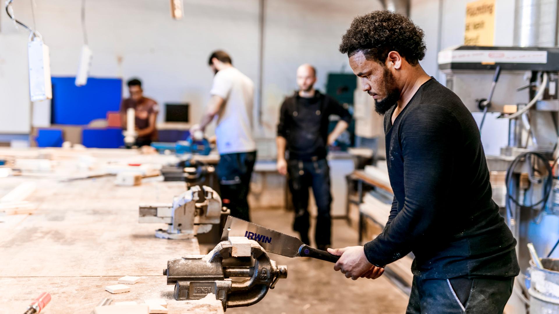 Ein Geflüchteter arbeitet in einer Holzwerkstatt und arbeitet an einem Holzstück. Im Hintergrund stehen Kollegen.