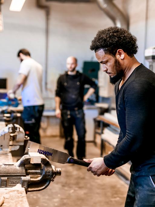 Ein Geflüchteter arbeitet in einer Holzwerkstatt und arbeitet an einem Holzstück. Im Hintergrund stehen Kollegen.