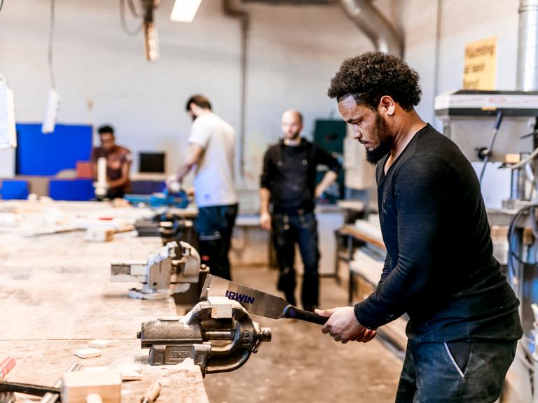 Ein Geflüchteter arbeitet in einer Holzwerkstatt und arbeitet an einem Holzstück. Im Hintergrund stehen Kollegen.