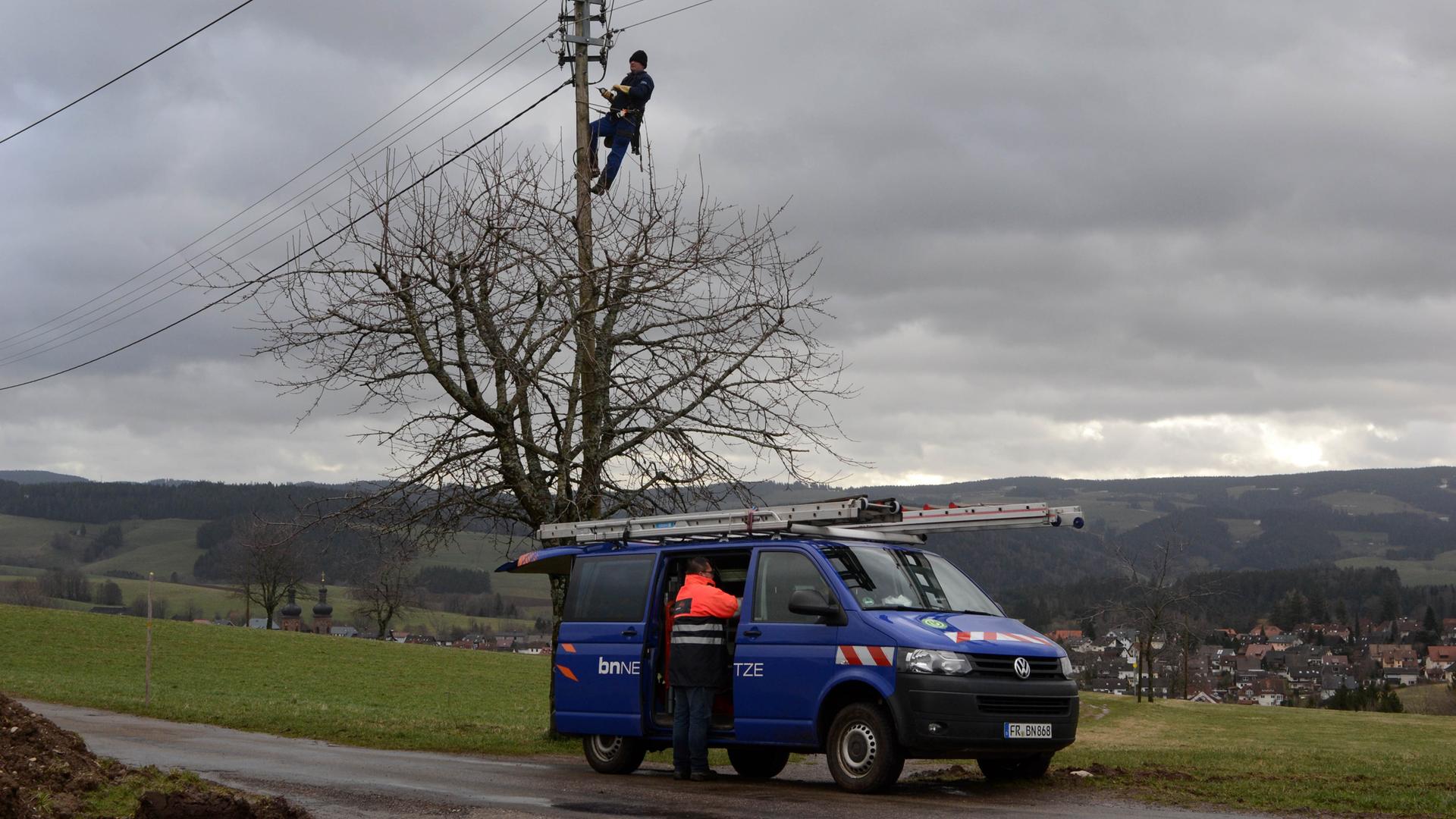 Am Rand einer Wiese ist ein Mann auf einen Strommast geklettert, davor steht ein Baum. Auf einem Feldweg steht ein blauer Wagen, in dem gerade ein anderer Mann etwas einräumt. Am Rand einer Wiese ist ein Mann auf einen Strommast geklettert, davor steht ein Baum. Auf einem Feldweg steht ein blauer Wagen, in dem gerade ein anderer Mann etwas einräumt.