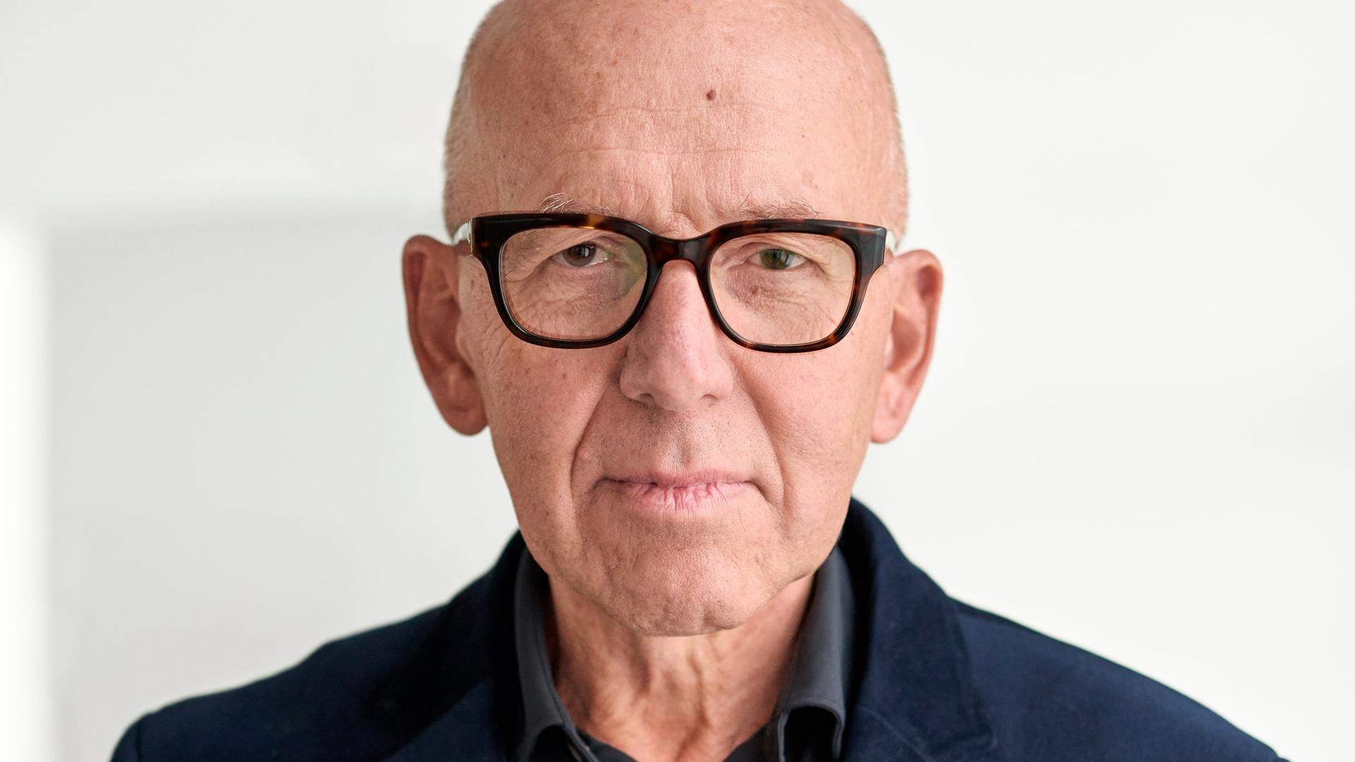 Ein Mann mit dunklem Hemd und Jackett und eckiger Hornbrille steht vor einer weißen Wand. Er schaut nachdenklich, frontal in die Kamera. Ein Mann mit dunklem Hemd und Jackett und eckiger Hornbrille steht vor einer weißen Wand. Er schaut nachdenklich, frontal in die Kamera.