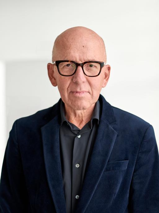 Ein Mann mit dunklem Hemd und Jackett und eckiger Hornbrille steht vor einer weißen Wand. Er schaut nachdenklich, frontal in die Kamera.