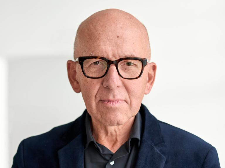 Ein Mann mit dunklem Hemd und Jackett und eckiger Hornbrille steht vor einer weißen Wand. Er schaut nachdenklich, frontal in die Kamera.