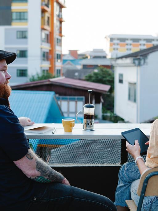 Zwei Menschen sitzen mit Getränken auf einem Balkon und unterhalten sich.