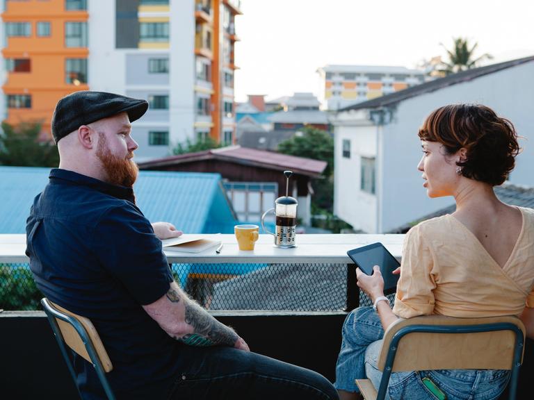 Zwei Menschen sitzen mit Getränken auf einem Balkon und unterhalten sich.